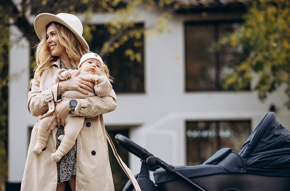 O femeie ține un bebeluș în brațe în timp ce merge pe lângă un cărucior în fața unei clădiri.